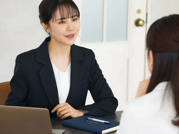 ダークスーツ姿の女性スタッフが、テーブル越しに向かい合うお客様へ笑顔で話しかけている打ち合わせの様子。テーブルの上にはノートパソコンと紺色の手帳、ペンが置かれ、和やかな雰囲気で相談が進められている。